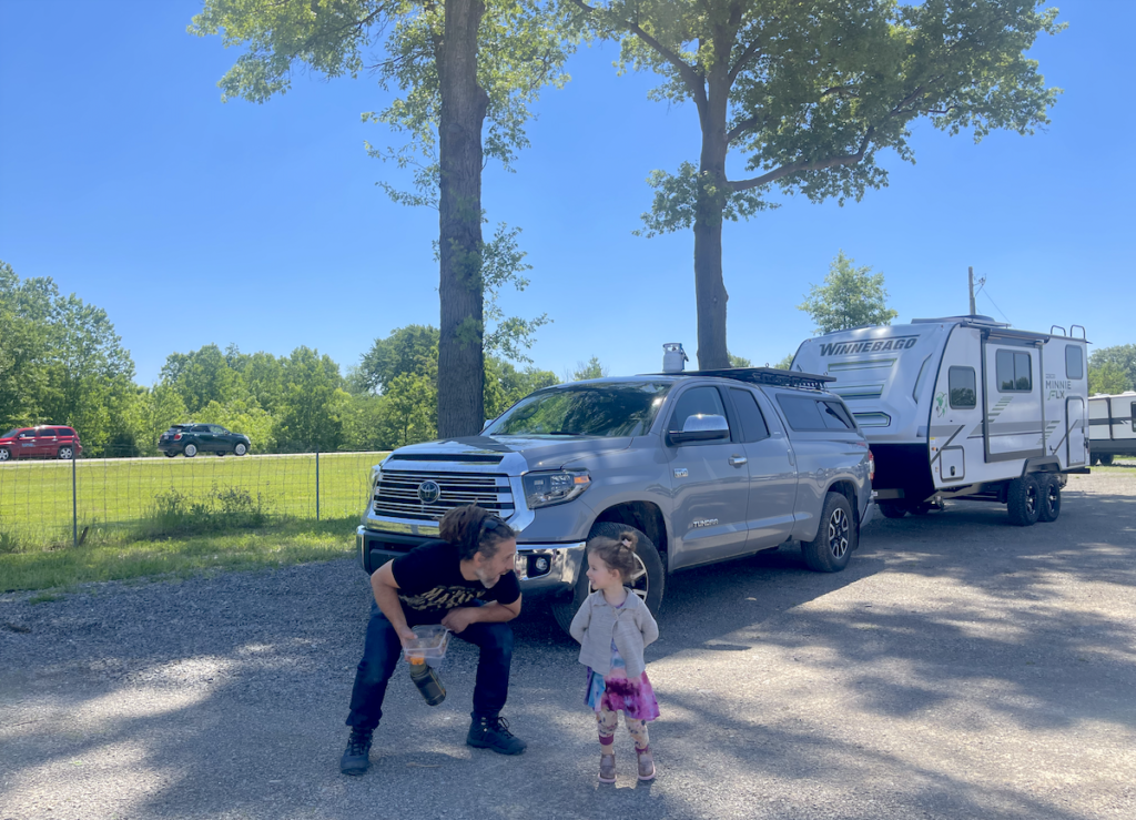 Man and girl stand outside an SUV towing a Winnebago travel trailer.