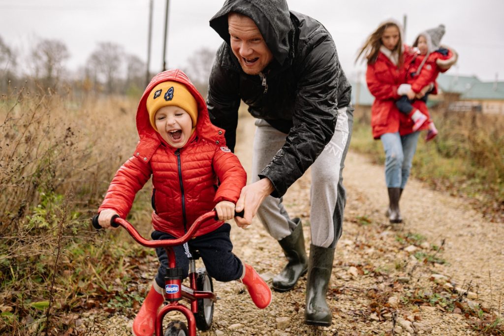 Dad pushes child on bicycle down dirt road while helping her balance. Mom and chlid in the background.