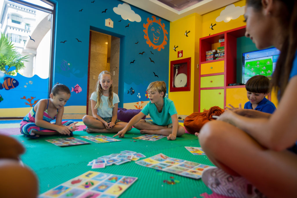 Four children are seated and playing a board game while a woman watches them.