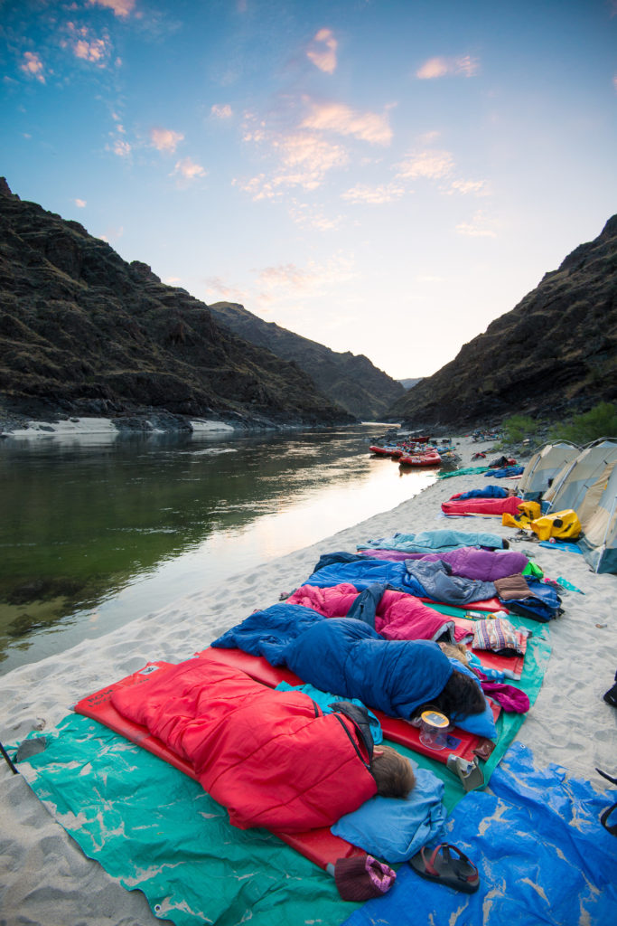 People in sleeping bags sleep out on the sand outside their tents by the side of the Salmon River.