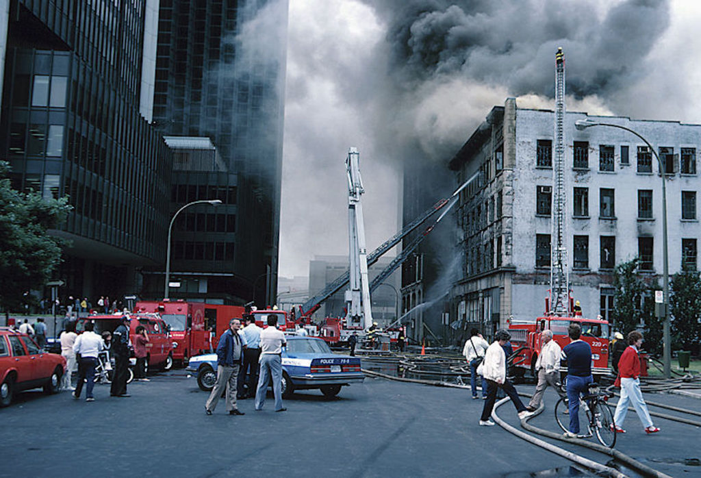 City street scene with firetrucks and police cars in front of a building with smoke coming out.