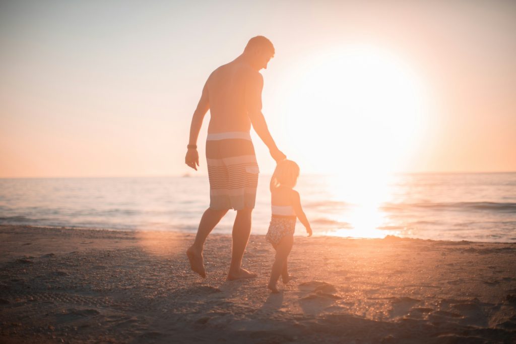 Parent supervising a young girl on the beach as she walks towards the water at dusk.