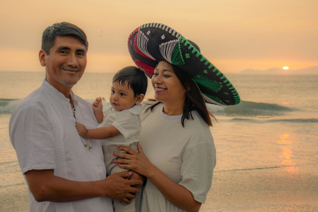Dad and mom in sombrero holding baby stand at the water's edge at dusik.