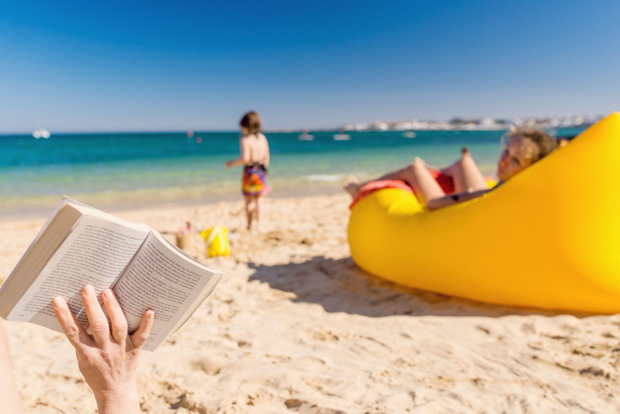 Mom reads book at beach while 2 toddlers play in sand nearby.